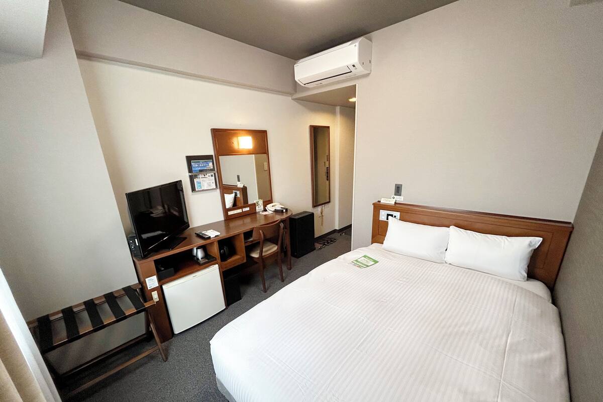 A hotel room with a bed, a desk with a chair, a television, a mirror, and an air conditioning unit.