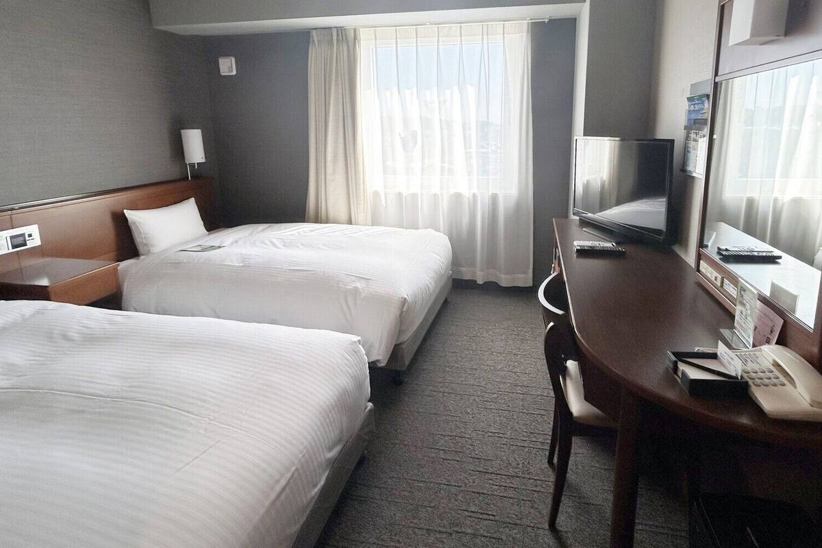 A hotel room with two beds, a desk with a phone, a TV, and a window with curtains.