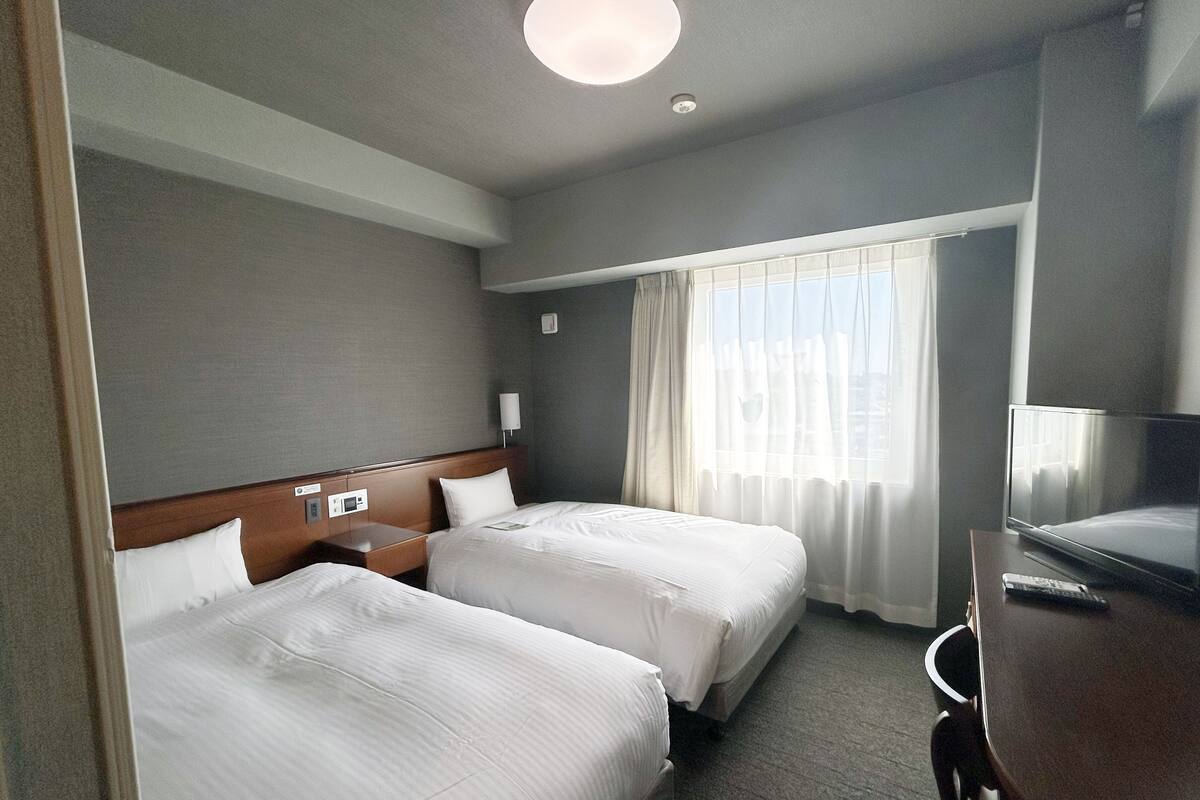 A hotel room with two beds, a desk, a television, and a window with curtains.
