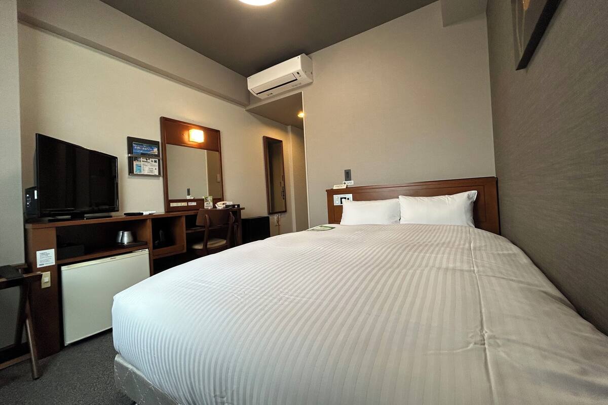 A hotel room with a bed, a desk with a computer, a television, and an air conditioning unit.