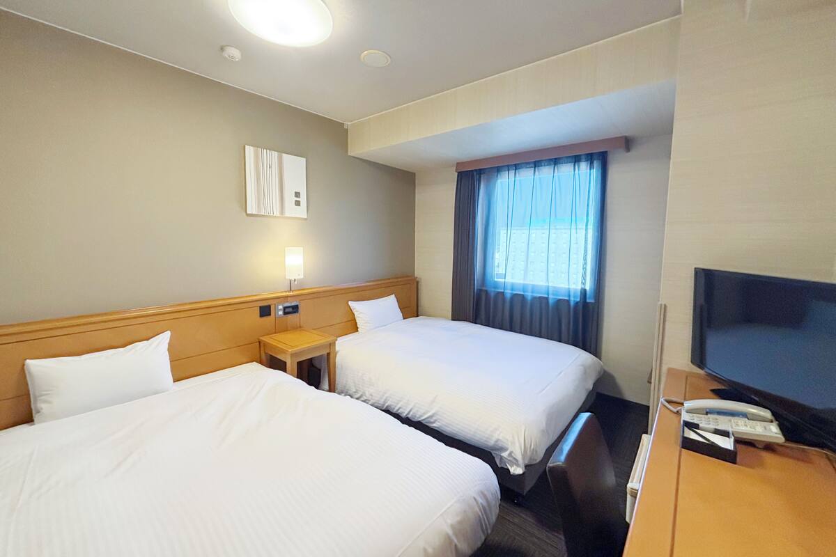 Quarto de hotel com duas camas, televisão, escrivaninha e janela com cortinas.