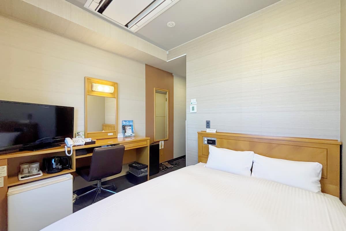 Quarto de hotel com cama, escrivaninha com computador, televisão, espelho e pequeno frigorífico.