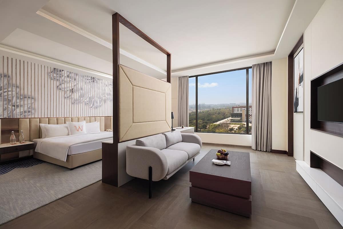 A modern hotel room with a bed, sofa, coffee table, and a large window offering a city view.
