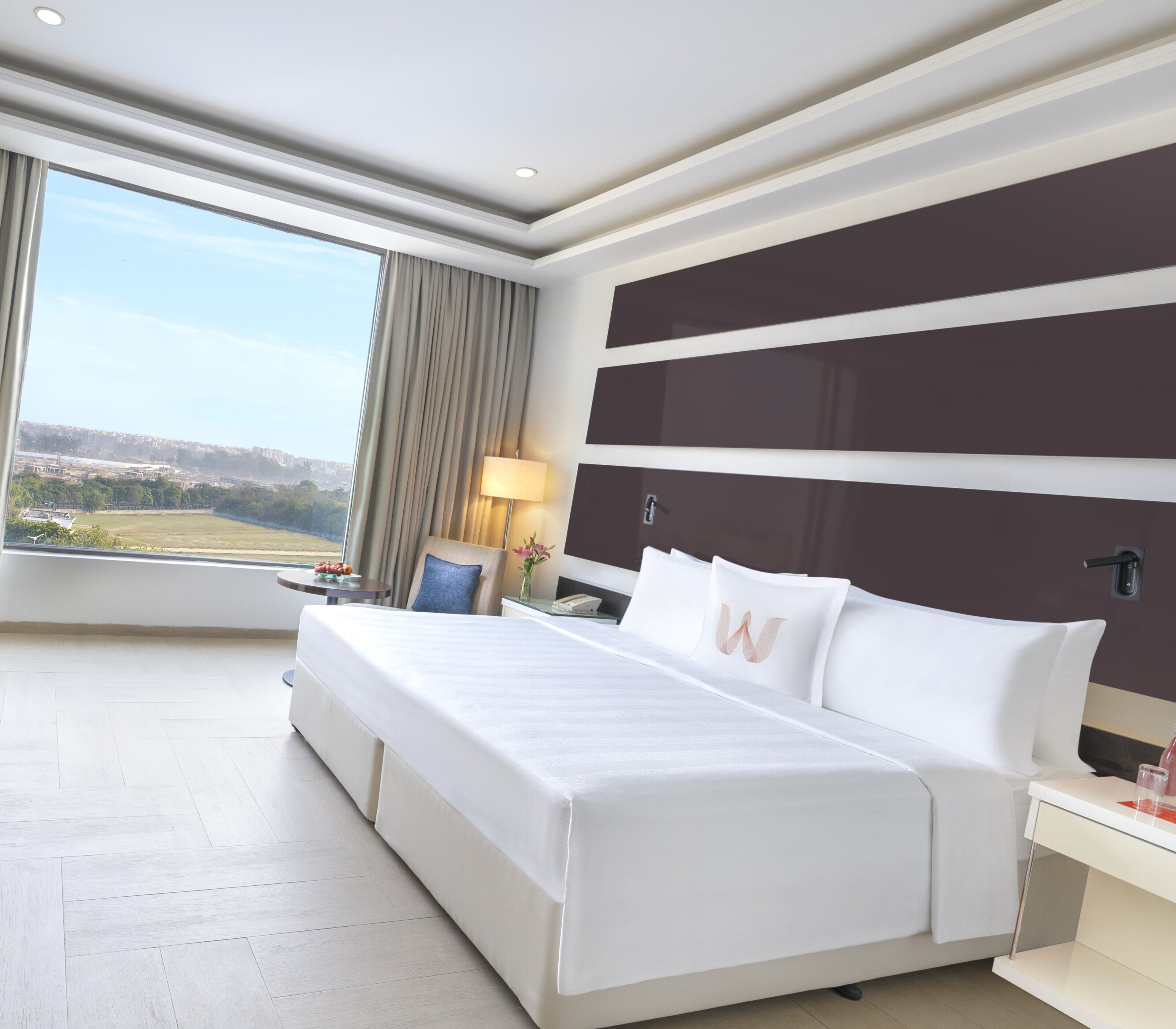 A modern hotel room with a large bed, a nightstand, a lamp, and a view of the cityscape.