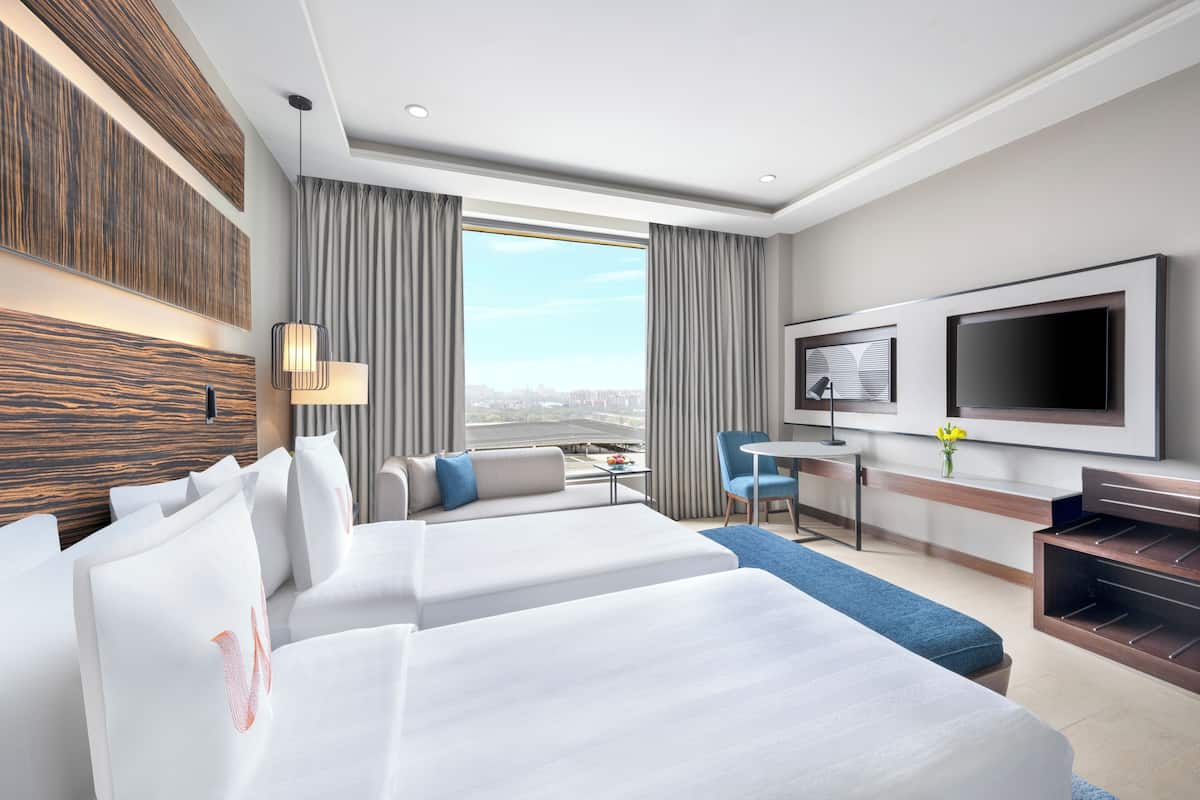 A hotel room with two beds, a TV, a desk, and a view of the city.