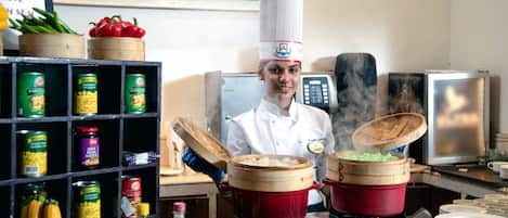 Un chef en una cocina con varias ollas, un estante con conservas y una pared decorada con cuadros.