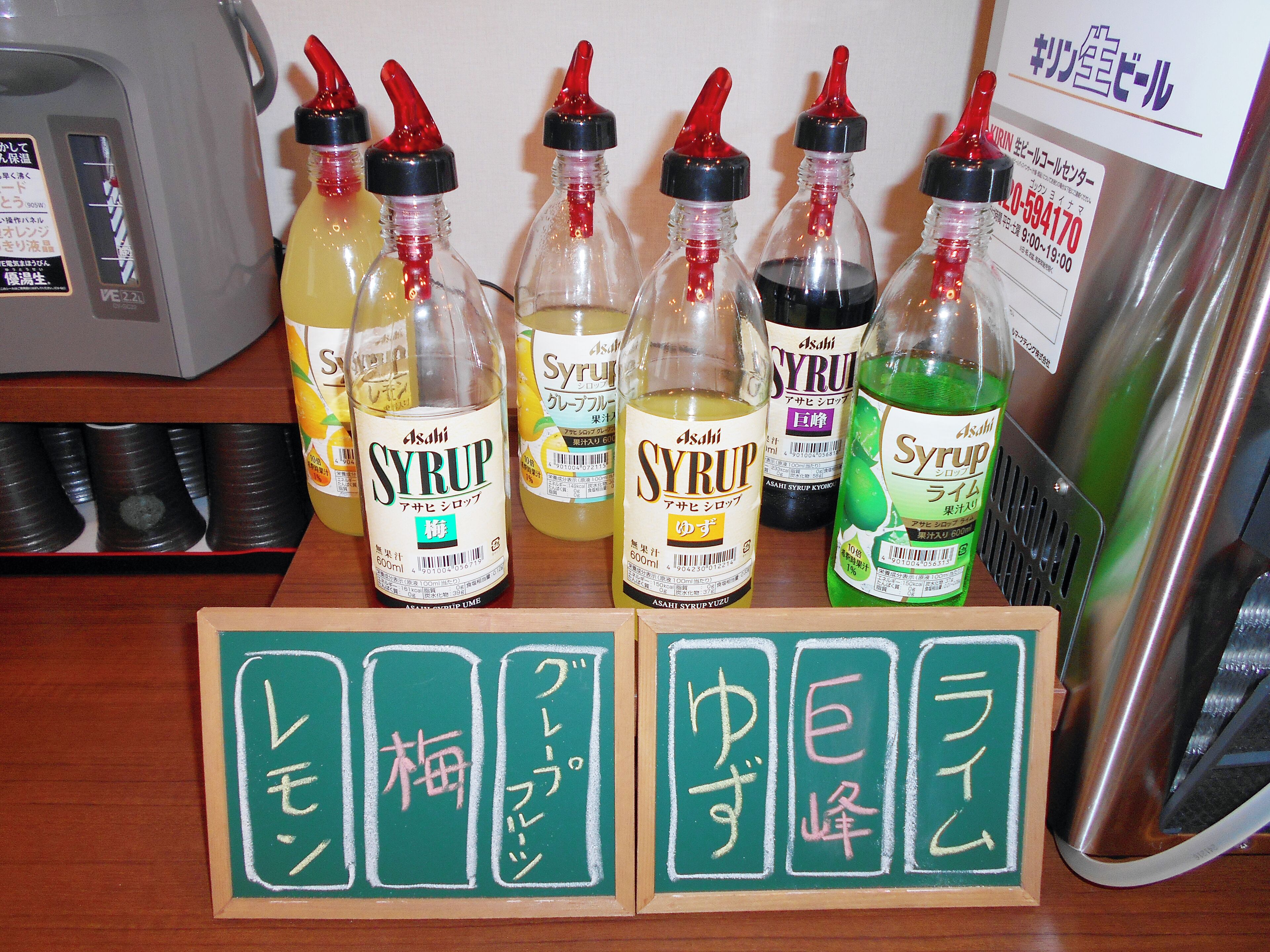 A display of Asahi Syrup bottles in various colors with labels, and a wooden board with Japanese characters.
