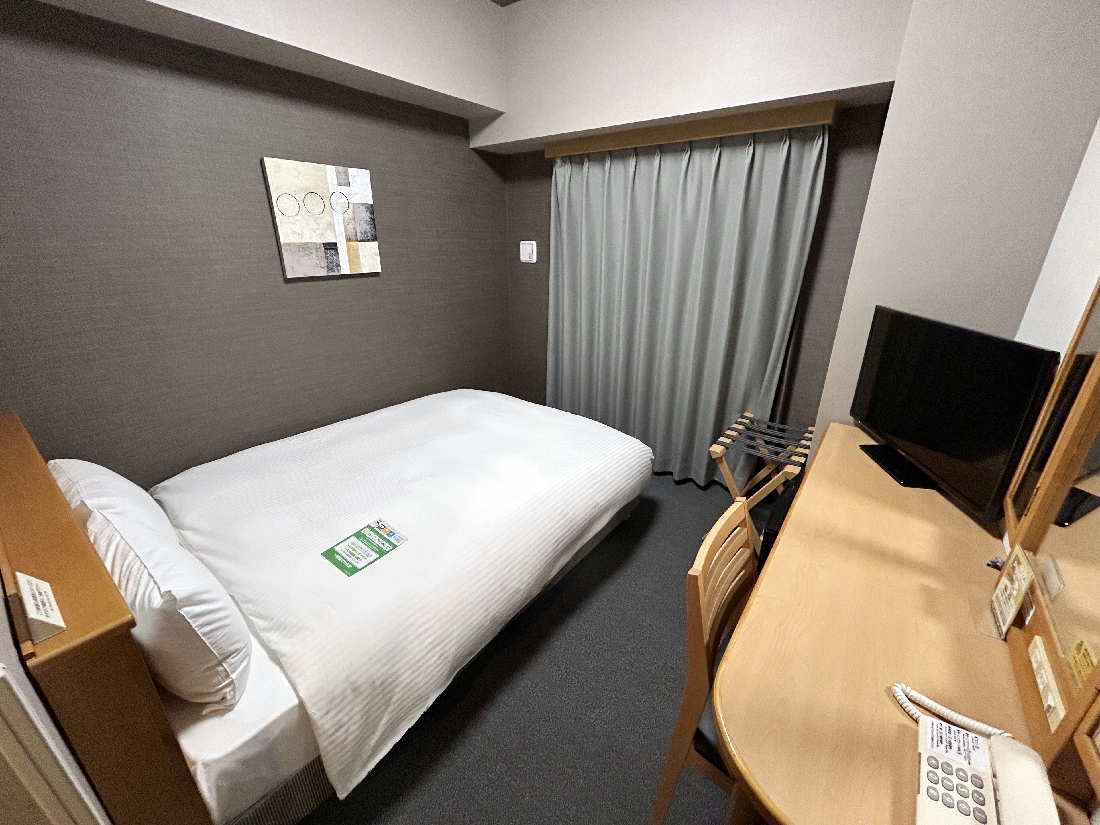A hotel room with a single bed, a desk with a telephone, a television, a window with curtains, and a wall-mounted artwork.