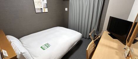 A hotel room with a single bed, a desk with a telephone, a television, a window with curtains, and a wall-mounted artwork.