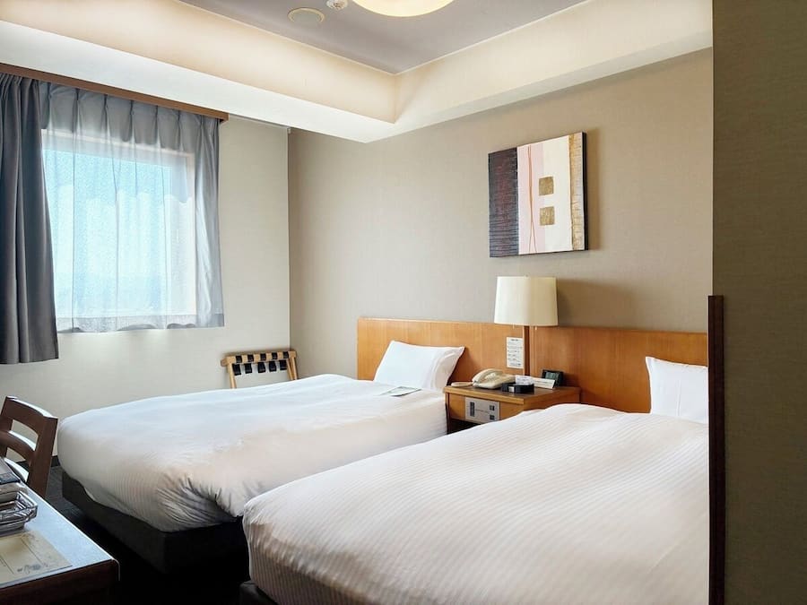 A hotel room with two beds, a desk, a lamp, and a painting on the wall.