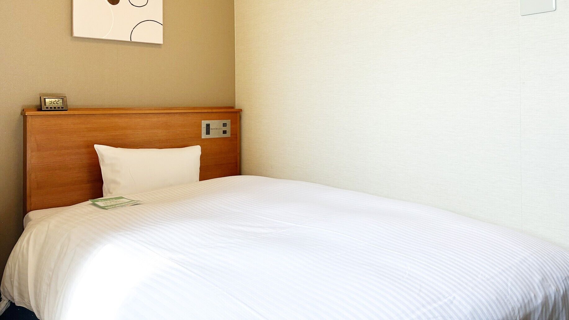 Quarto de hotel com cabeceira de madeira, colcha branca, travesseiro branco, mesa de cabeceira com relógio e interruptor de luz na parede.