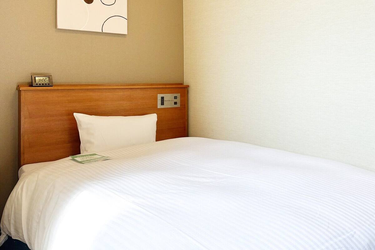 Ein Hotelzimmer mit hölzernem Kopfteil, weißer Bettdecke, einem weißen Kissen, einem Nachttisch mit einer Uhr und einem an der Wand befestigten Lichtschalter.
