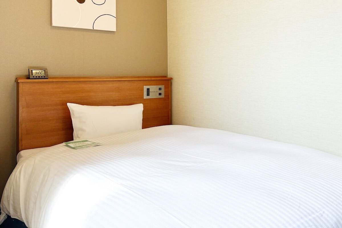Quarto de hotel com cabeceira de madeira, colcha branca, travesseiro branco, mesa de cabeceira com relógio e interruptor de luz na parede.