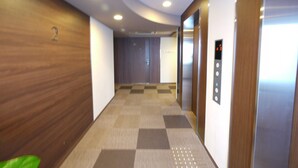 A hallway with wooden walls, a checkered floor, and an elevator with a digital display. - Hotel Route Inn Ishinomaki Kanan Inter (Isinomaki)