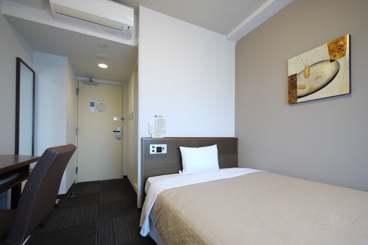 Habitación de hotel con cama, escritorio y silla. En la pared hay un cuadro y una puerta con un cartel.