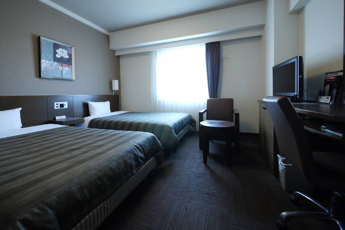 Habitación de hotel con dos camas, un escritorio con silla, televisión y ventana con cortinas.