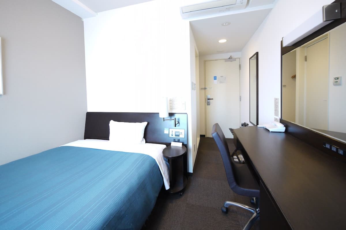 Una habitación de hotel con cama, escritorio, silla, espejo y aire acondicionado.