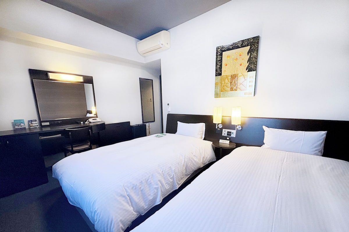 A hotel room with two beds, a desk, a chair, a TV, and a wall-mounted air conditioner.