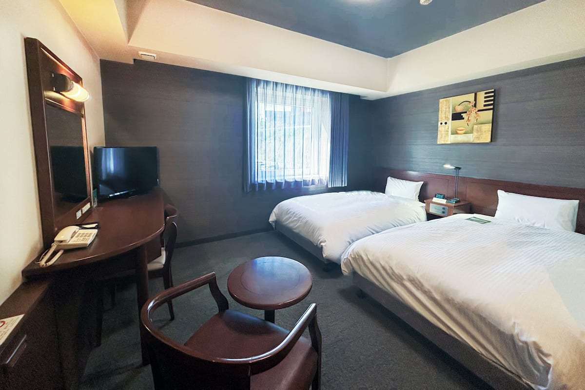 A hotel room with two beds, a desk with a chair, a television, and a window with curtains.