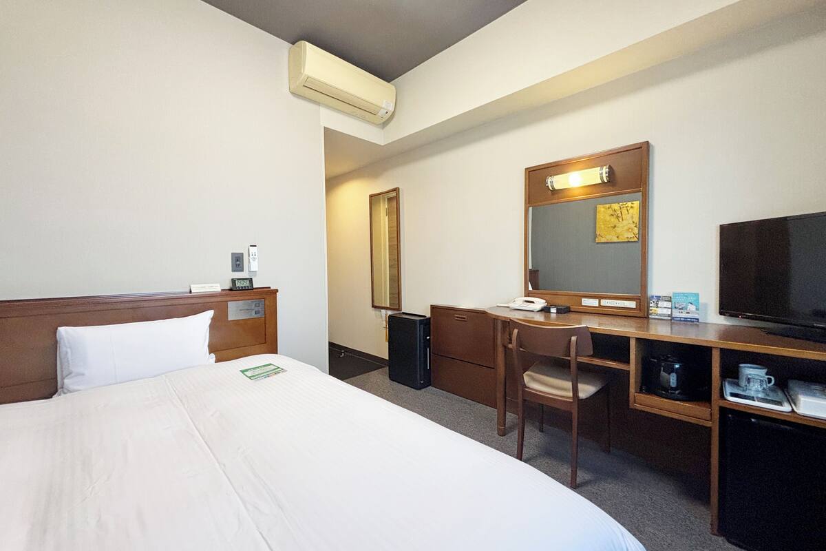 A hotel room with a bed, a desk, a chair, a television, a mirror, and an air conditioning unit.
