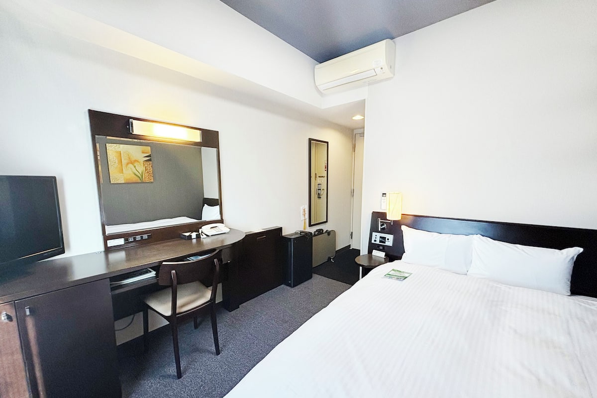 A hotel room with a bed, a desk, a chair, a TV, a mirror, a bedside table, and an air conditioning unit.