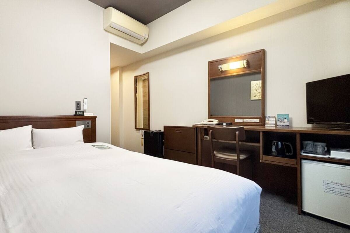 A hotel room with a large bed, a desk with a chair, a television, a mirror, and a small refrigerator.