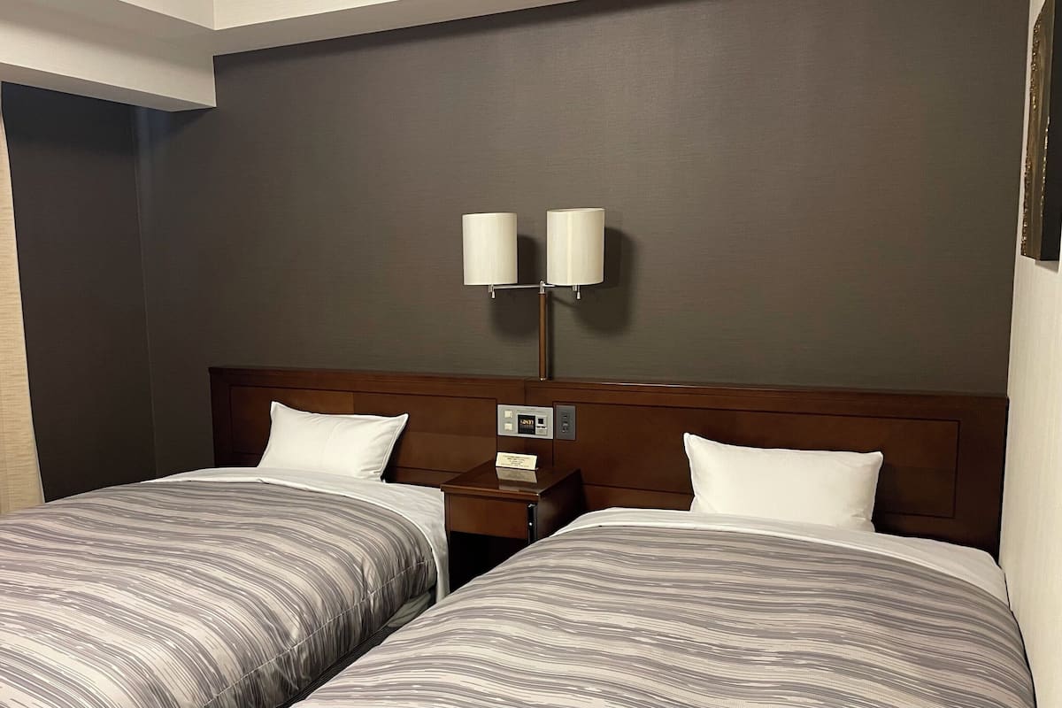 A hotel room with two beds, a nightstand, a wall-mounted lamp, and a window with curtains.