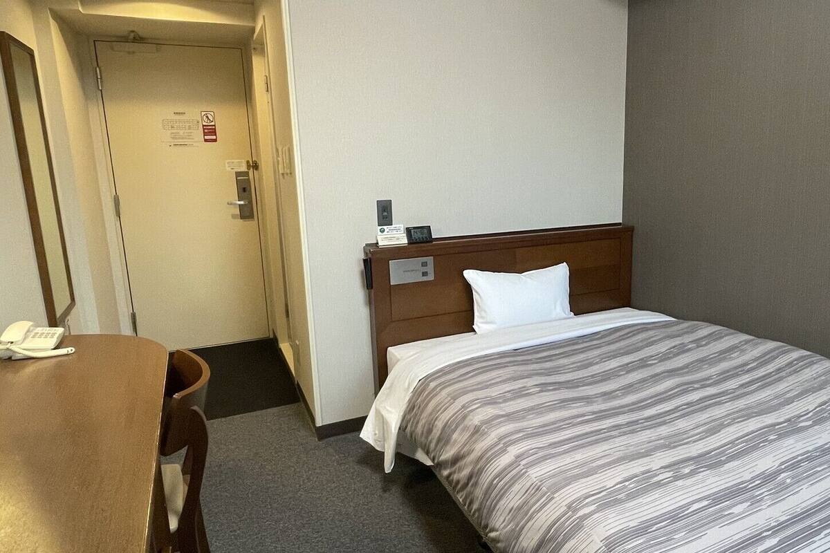 Quarto de hotel com cama, escrivaninha, telefone e porta.