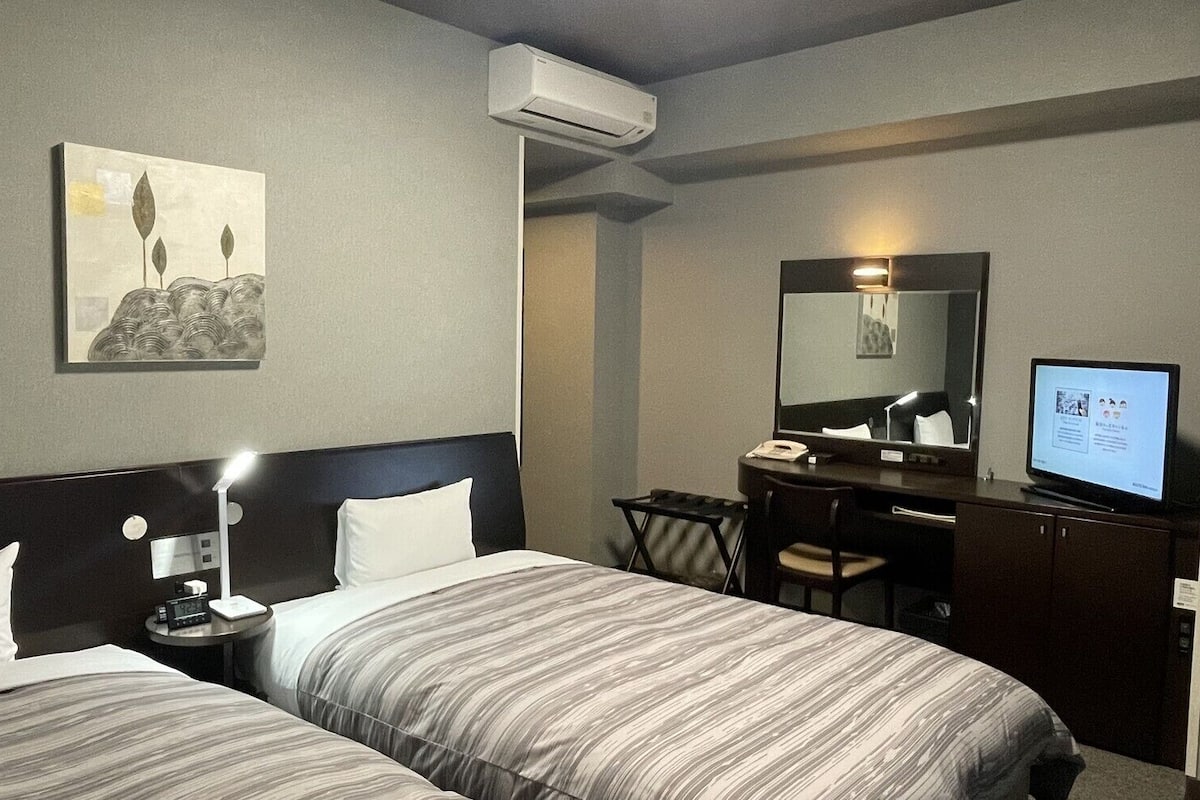 Quarto de hotel com duas camas, uma mesa com cadeira, televisão e espelho.
