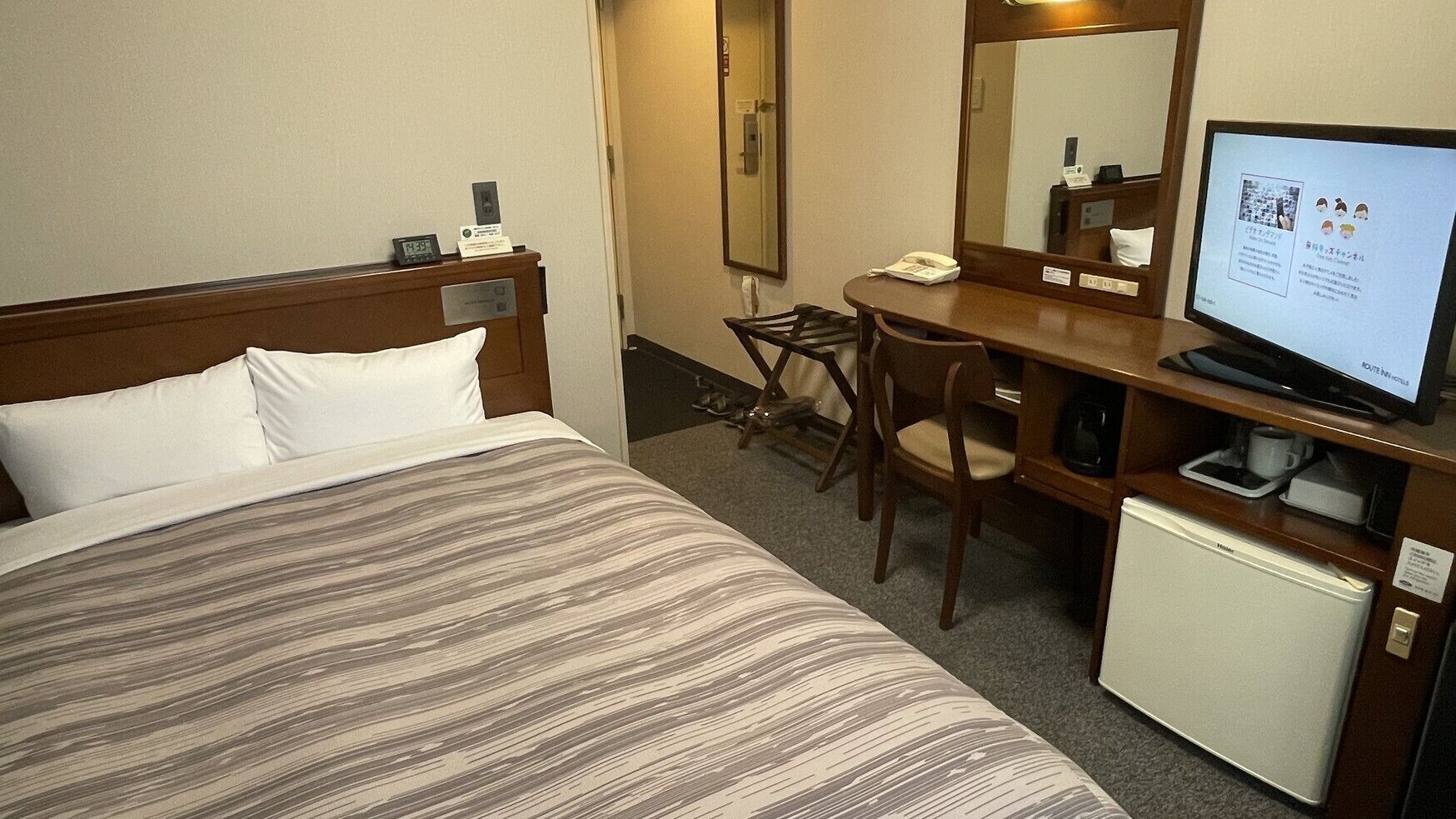 Quarto de hotel com cama, escrivaninha, cadeira, televisão e um pequeno frigorífico.