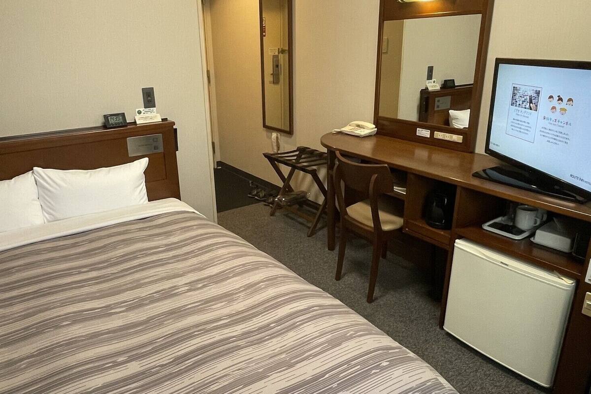Quarto de hotel com cama, escrivaninha, cadeira, televisão e um pequeno frigorífico.