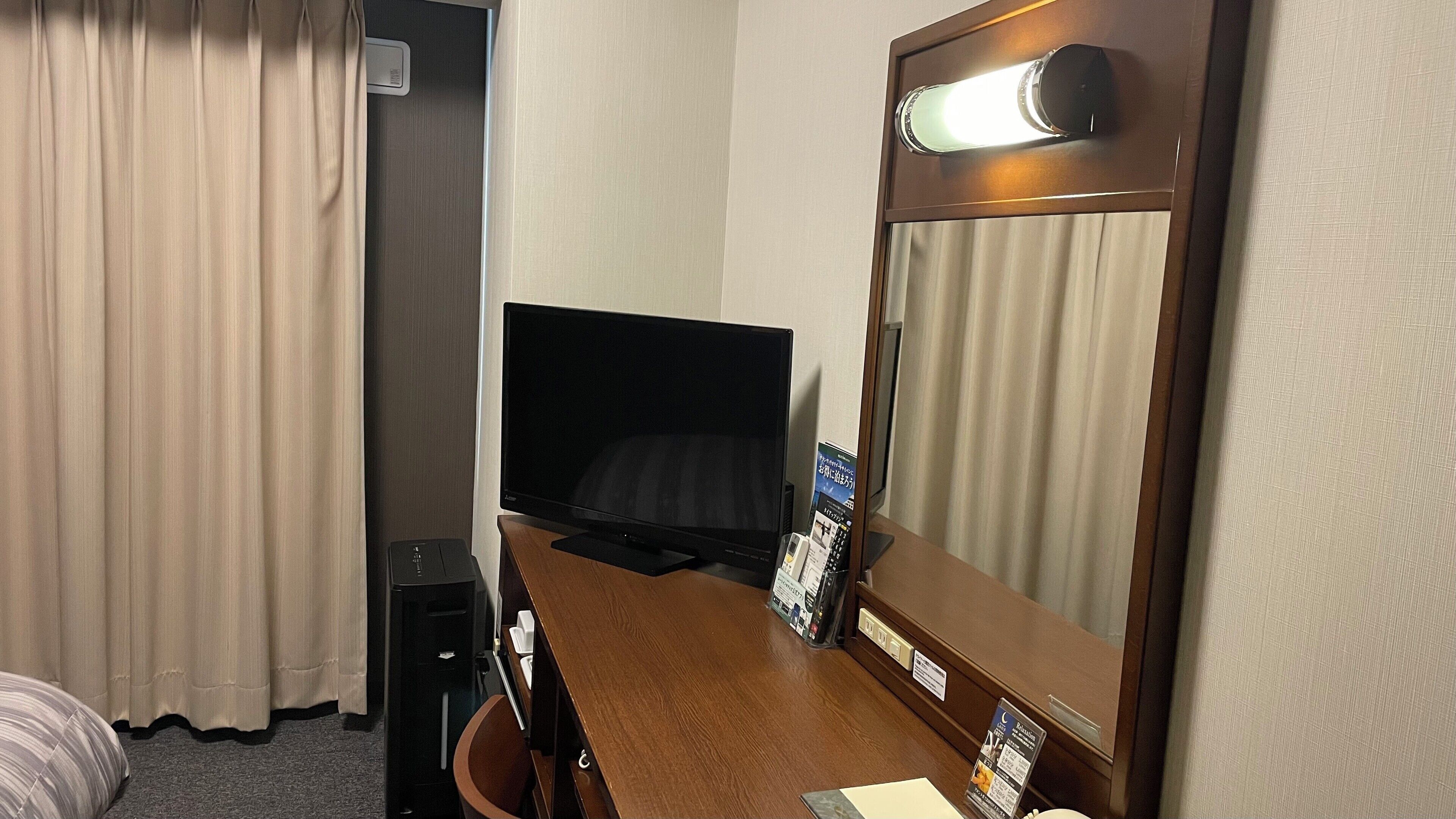 Quarto de hotel com cama, escrivaninha com computador, televisão, espelho e janela com cortinas.