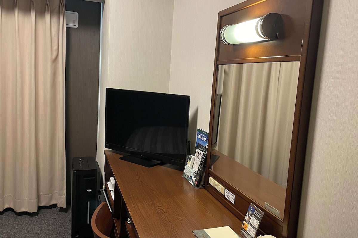 Quarto de hotel com cama, escrivaninha com computador, televisão, espelho e janela com cortinas.