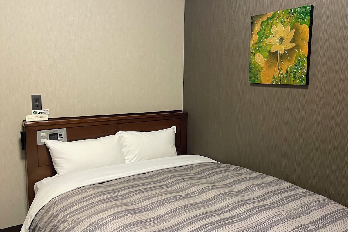 Quarto de hotel com cama, cabeceira, obra de arte na parede e painel de controle.