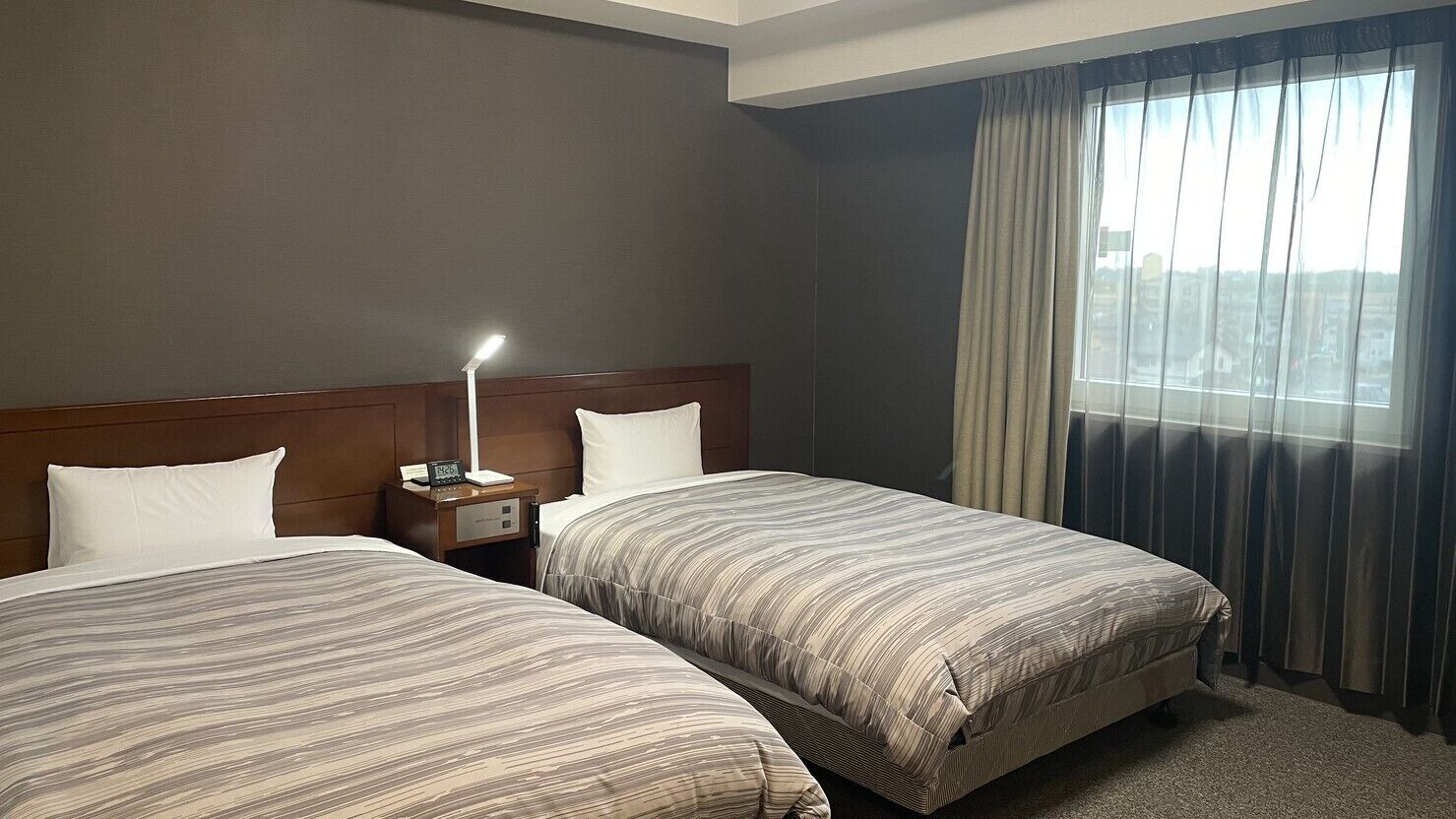 Quarto de hotel com duas camas, uma mesa de cabeceira com abajur, uma janela com cortinas e vista para edifícios.