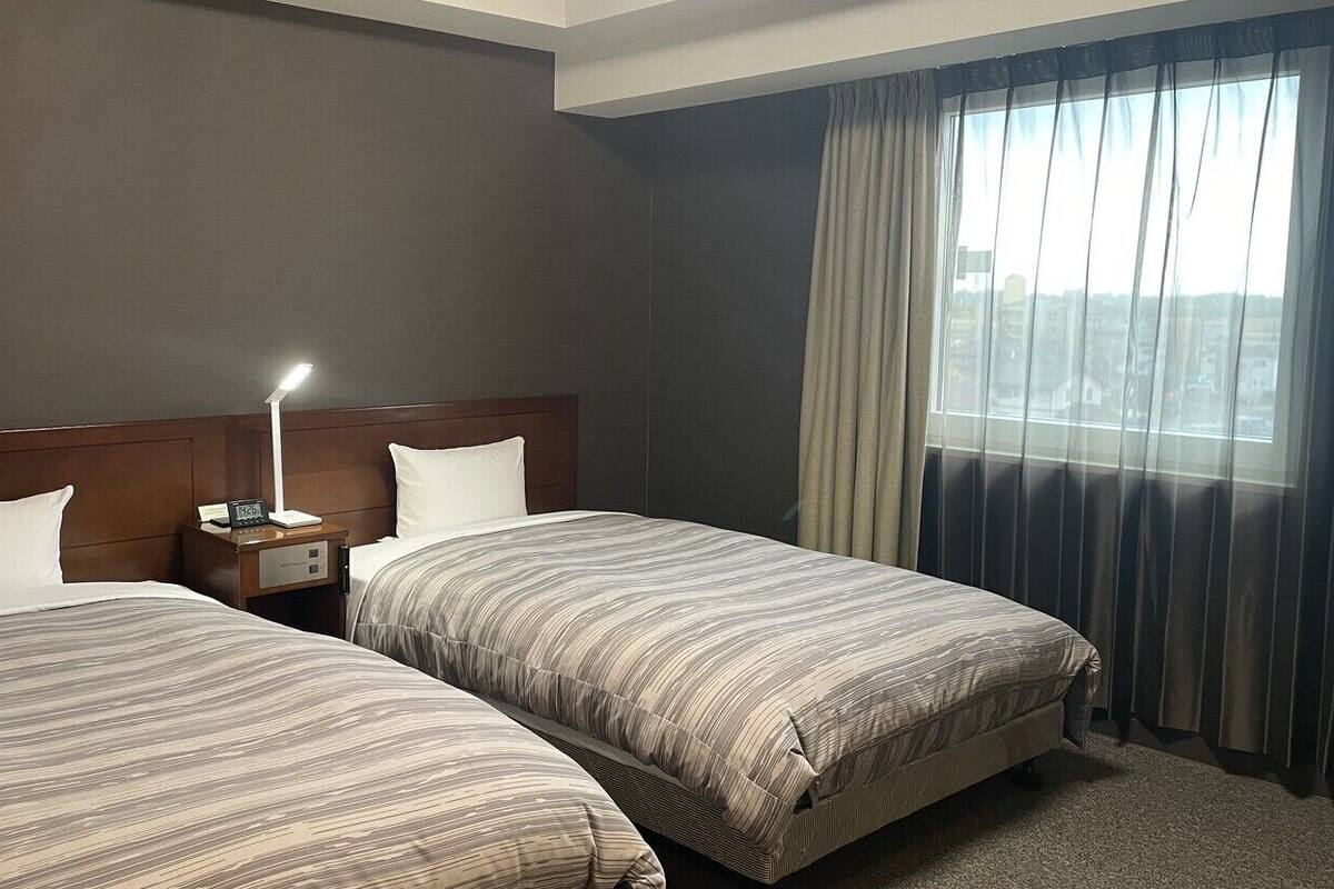 Quarto de hotel com duas camas, uma mesa de cabeceira com abajur, uma janela com cortinas e vista para edifícios.