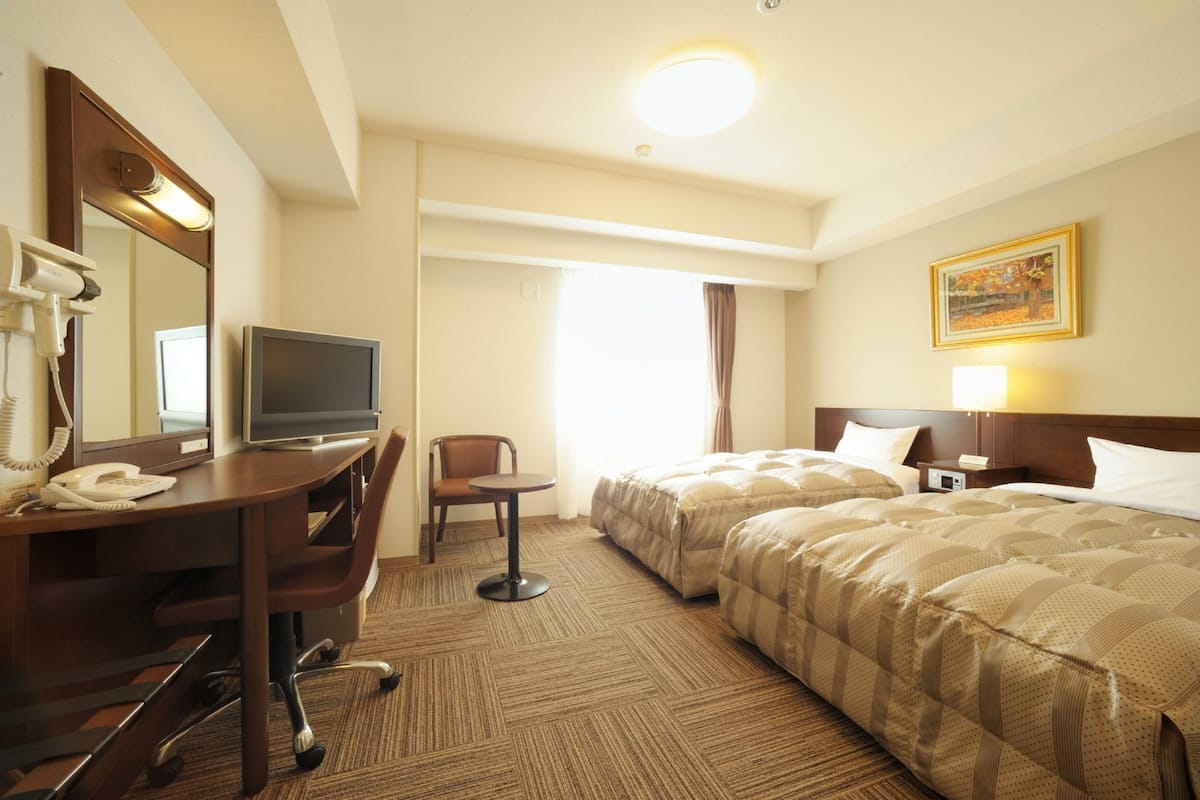 A hotel room with a desk, chair, television, two beds, and a large window.