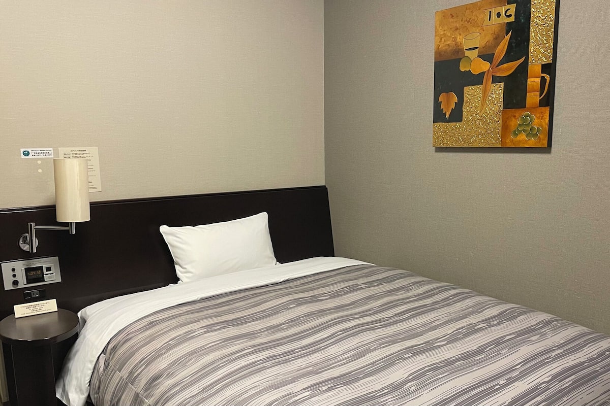 Quarto de hotel com uma cama, mesa de cabeceira, luminária na parede e um quadro.