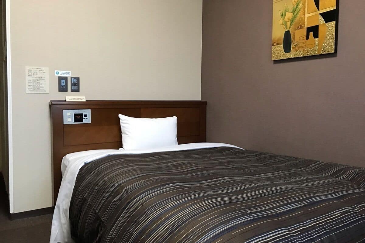 A hotel room with a bed, a control panel, a wall-mounted artwork, and a door.
