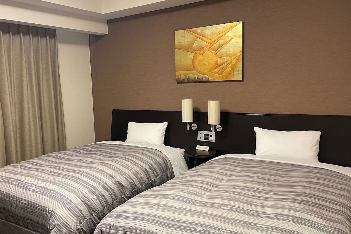 Quarto de hotel com duas camas, uma janela grande com cortinas, uma obra de arte na parede e luminárias de cabeceira.