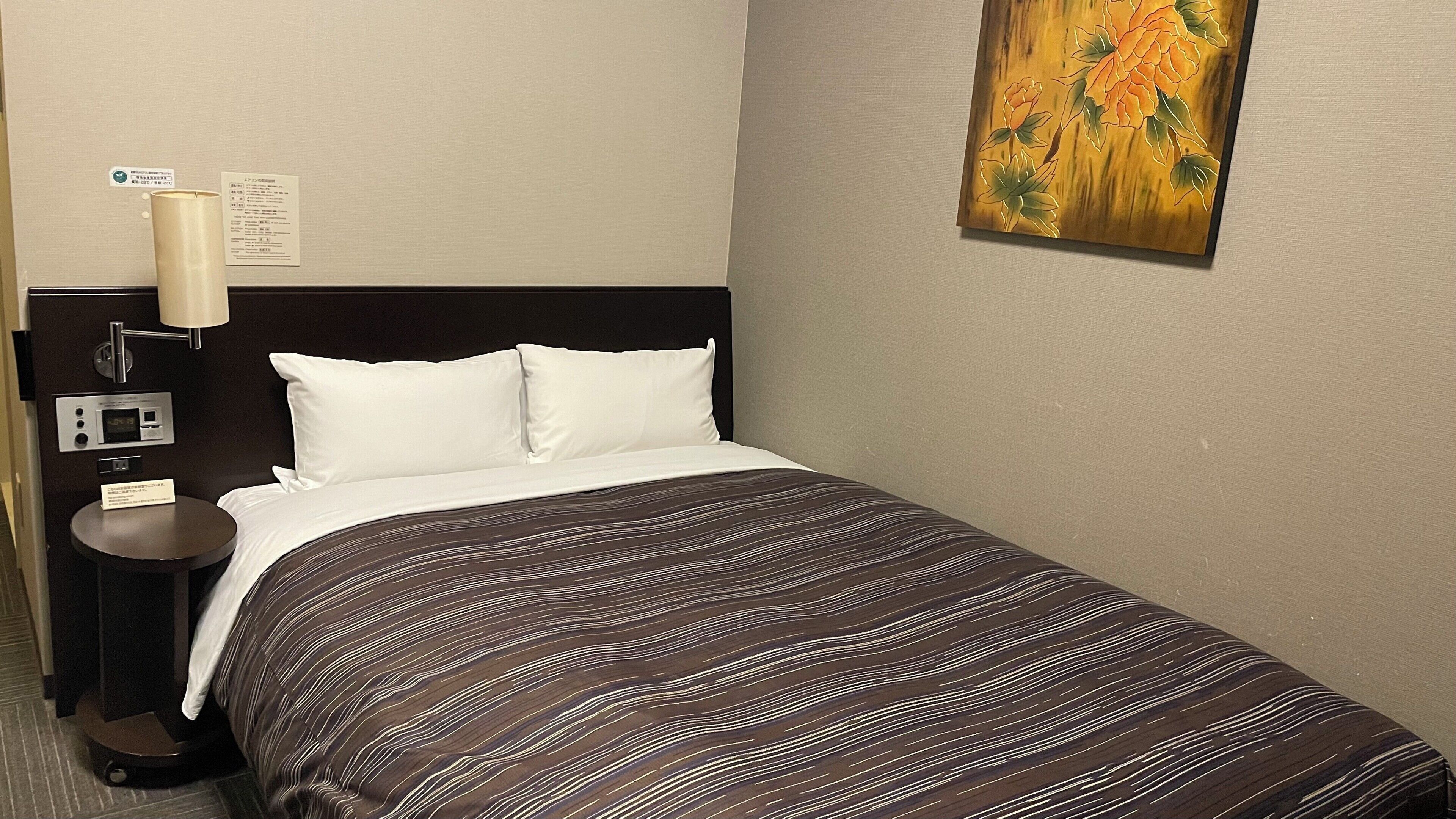 Quarto de hotel com cama, mesa de cabeceira, abajur, painel de controle e um quadro na parede.