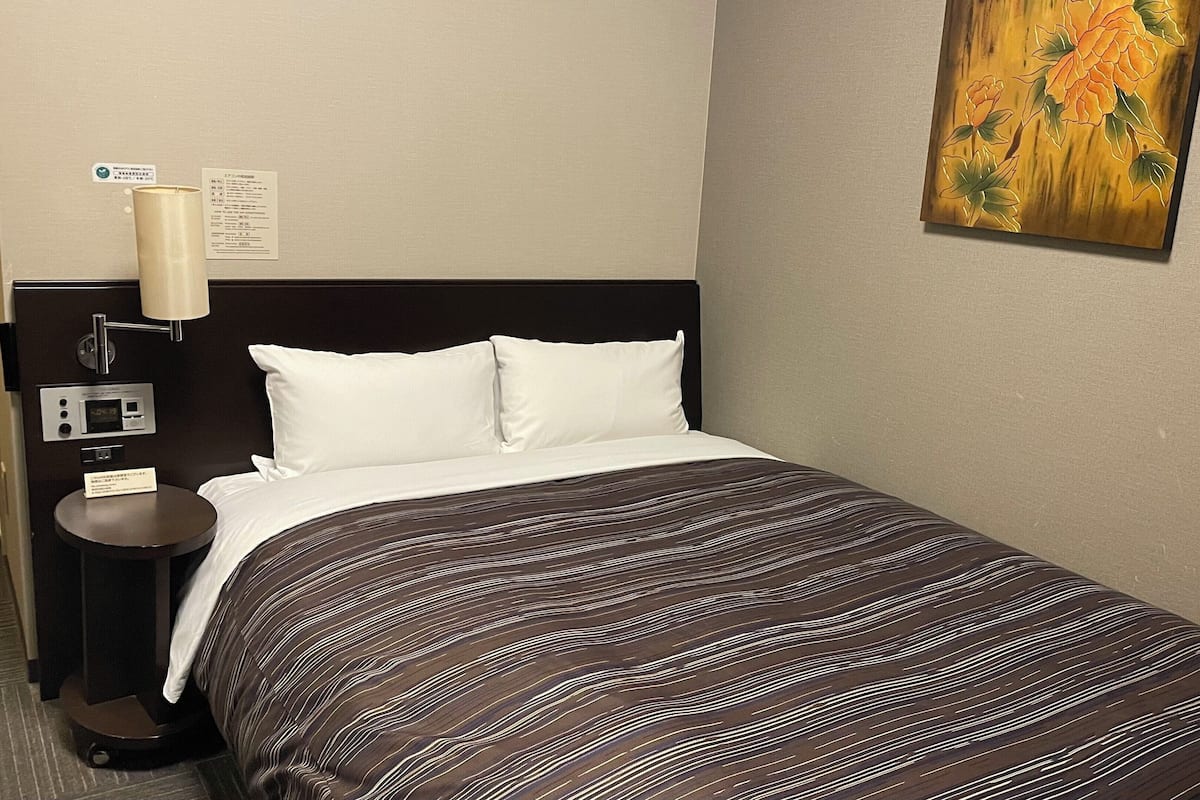 Quarto de hotel com cama, mesa de cabeceira, abajur, painel de controle e um quadro na parede.
