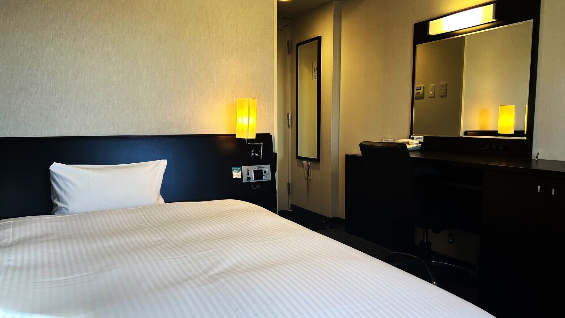 A hotel room with a bed, bedside table, lamp, mirror, and a desk with a telephone.