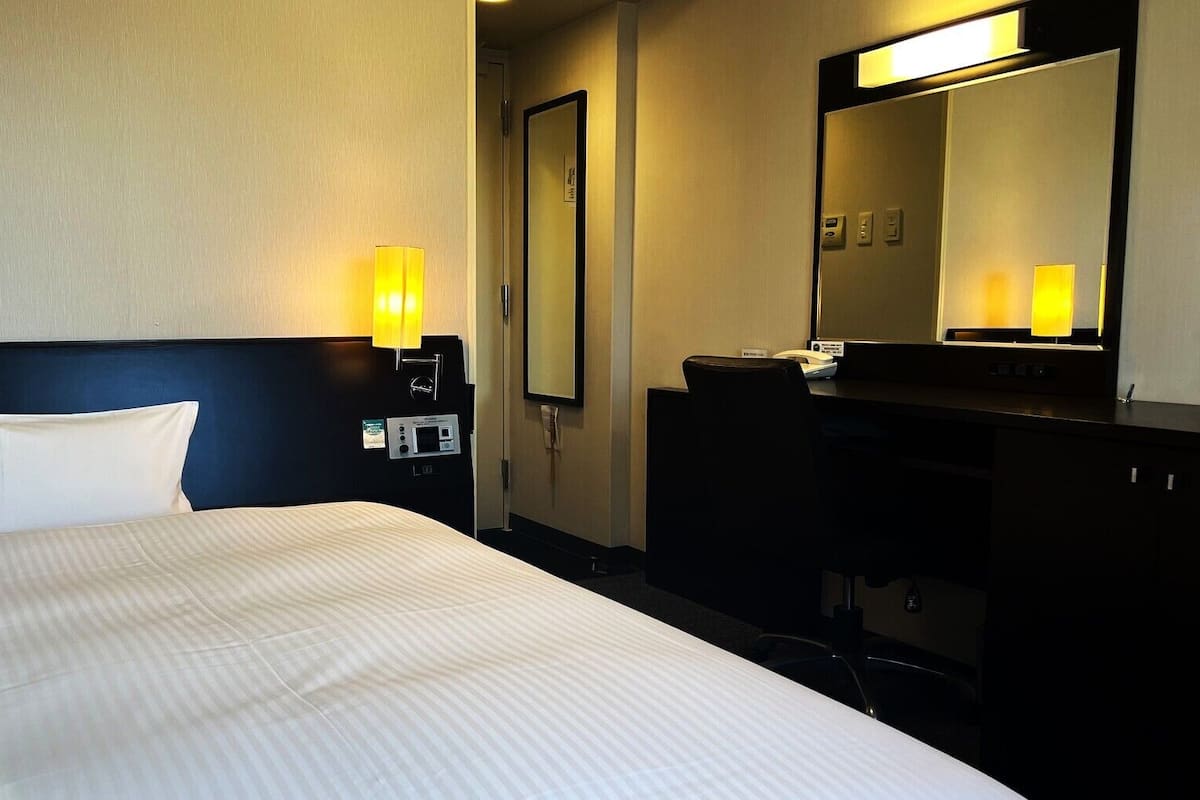 Habitación de hotel con cama, mesita de noche, lámpara, espejo y escritorio con teléfono.