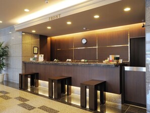 A reception area with a 'FRONT' sign, a clock, and a counter with a phone and informational brochures. There are three stools in front of the counter. - Hotel Route-Inn Sendaiko Kita Inter (Tagajo)