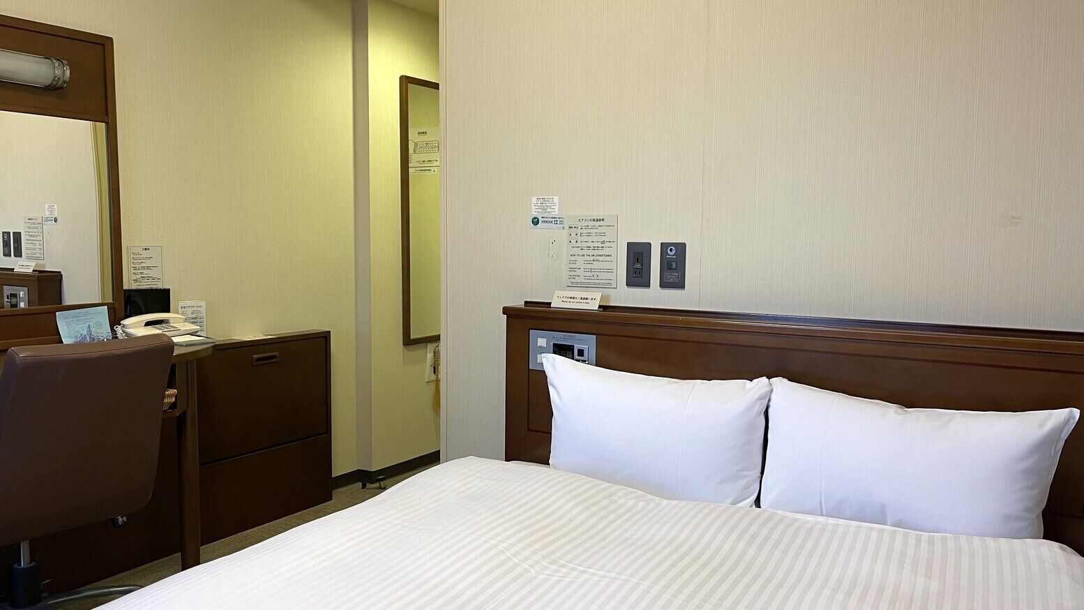 A hotel room with a bed, a desk with a telephone, a mirror, and a wall with informational signs.