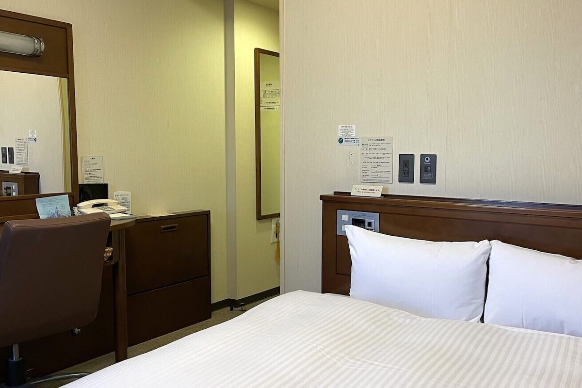 Habitación de hotel con una cama, un escritorio con teléfono, un espejo y una pared con carteles informativos.