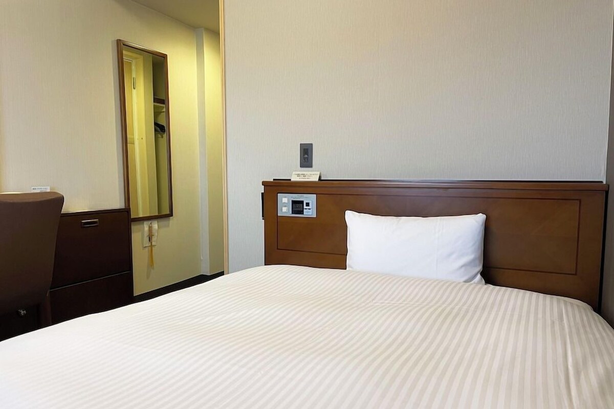 A hotel room with a large bed, a wooden headboard, a bedside table, a mirror, and a wall-mounted control panel.