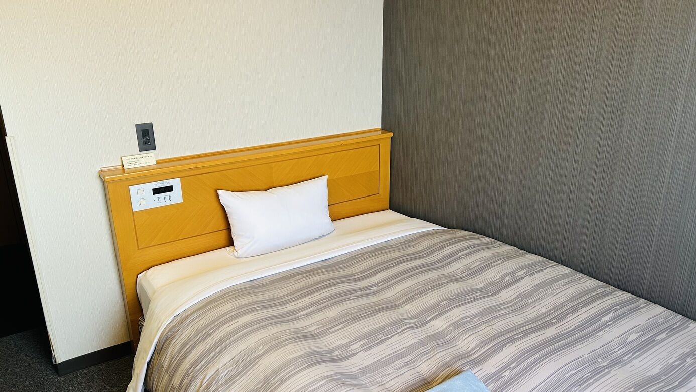 A hotel room with a wooden headboard, a single white pillow, a striped bedspread, and a wall-mounted control panel.