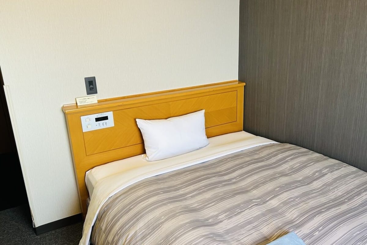 Ein Hotelzimmer mit einem hölzernen Kopfteil, einem weißen Kissen, einer gestreiften Tagesdecke und einer an der Wand befestigten Steuerung.